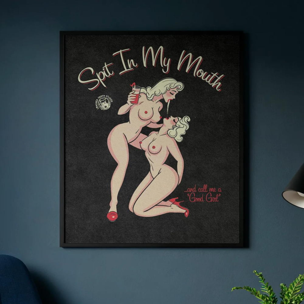 a cozy living room with a blue armchair, a wooden coffee table, and a framed poster on the wall. The poster depicts two naked women and includes the text "Spit In My Mouth" and "and all I want is a Good Old Club".