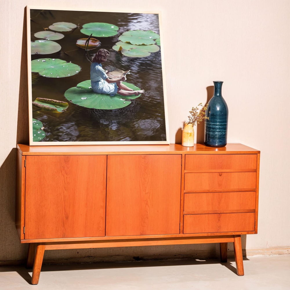 a wooden cabinet with a framed photograph of a person sitting on a lily pad in a pond, surrounded by green lily pads. The photograph is displayed on the cabinet, which is positioned against a wall.