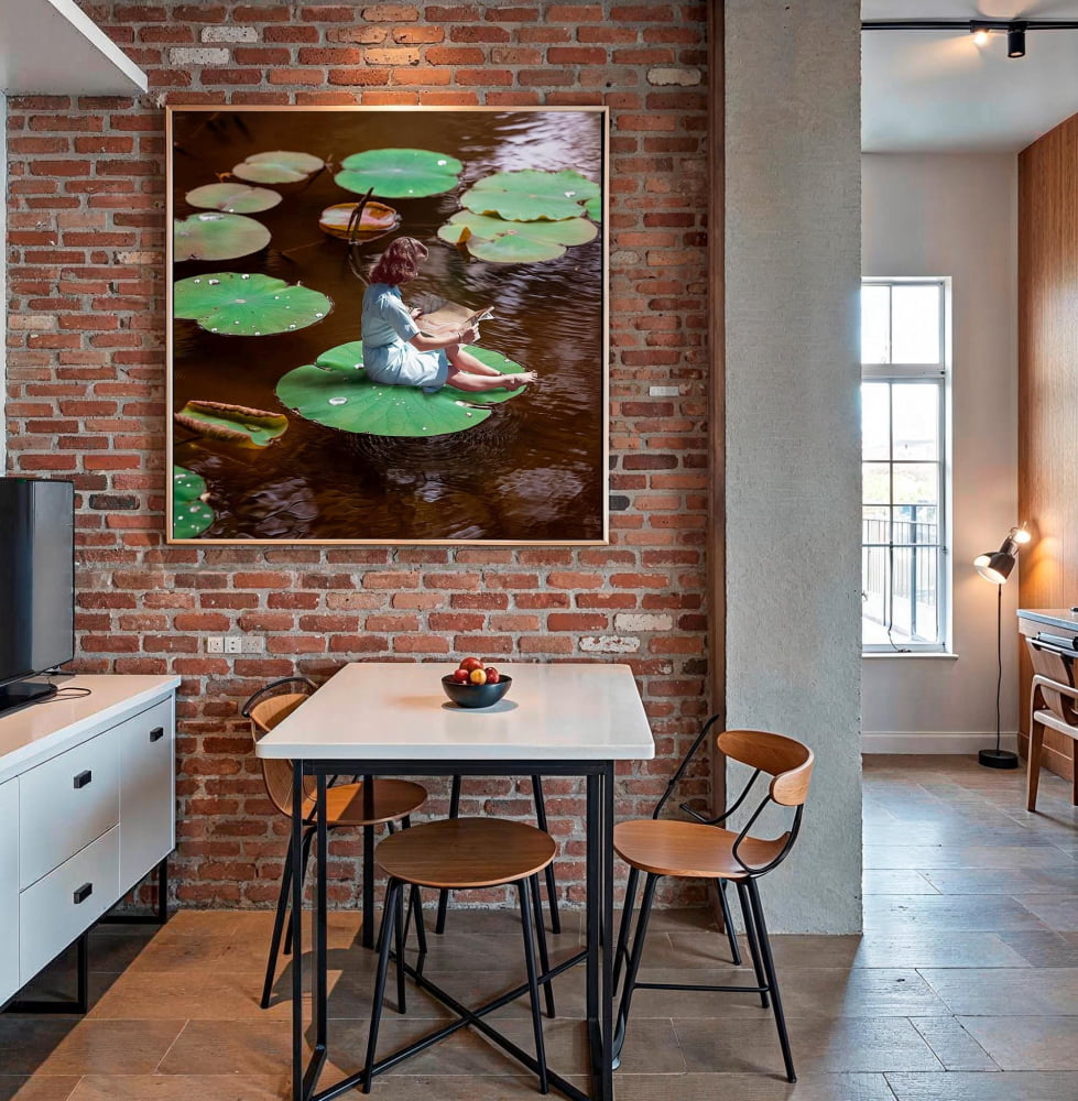 a dining area with a table and chairs, a large framed picture of a woman sitting on a lily pad, and a brick wall with a window.