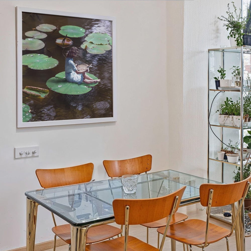 a glass dining table with four wooden chairs, and a framed painting of a person sitting on a lily pad above the table.