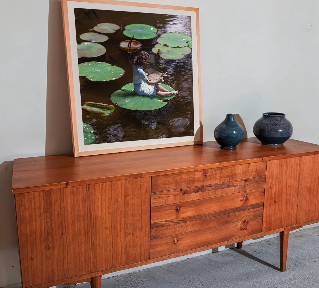 a wooden cabinet with a framed picture of a woman sitting on a lily pad in a pond, surrounded by green lily pads. The cabinet is placed against a white wall, and there are two blue vases on top of it.