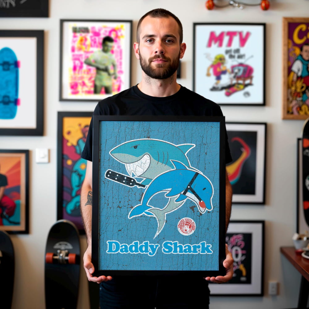 A man is holding a framed picture of a blue shark with a red tag on its fin, which says "Daddy Shark". The background features various framed pictures and artwork on the wall.