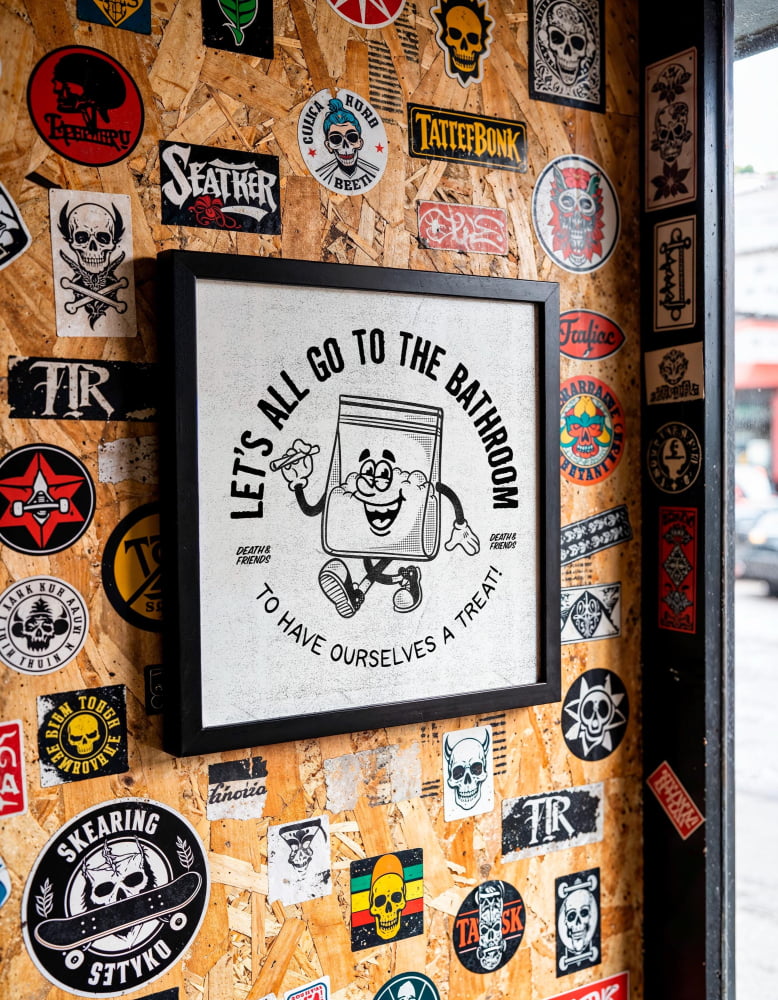 a wall covered in various stickers, posters, and graffiti, including a framed cartoon character with the text "Let's all go to the bathroom to have ourselves a treat!"