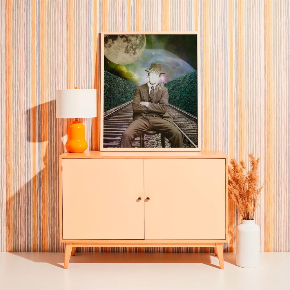 a framed artwork depicting a man sitting on a train track with a large moon in the background. The artwork is displayed on a wooden cabinet, which is positioned next to a lamp and a vase.