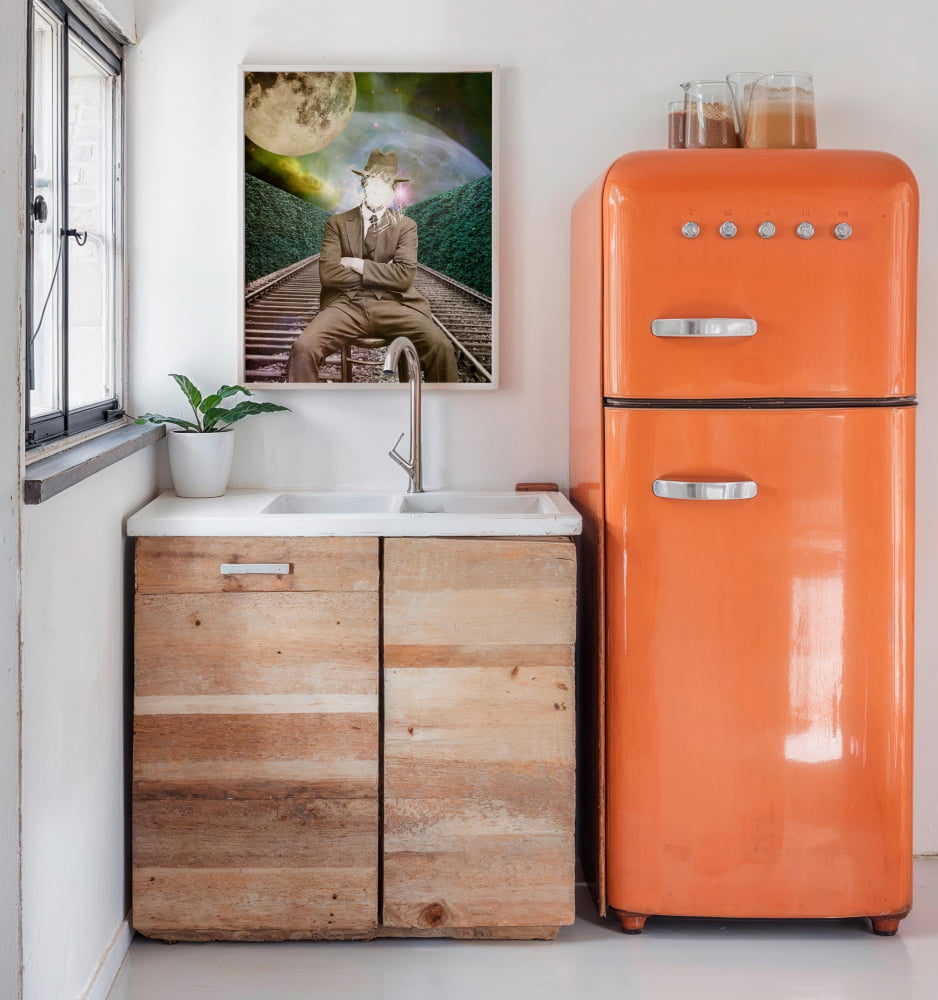 a kitchen scene with a wooden cabinet, a sink, and an orange retro-style refrigerator. Above the sink, there is a framed image of a man sitting on a train track.