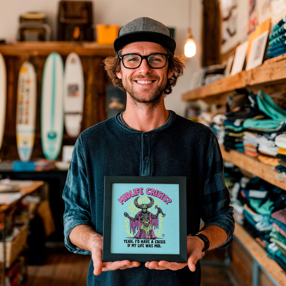 A man is holding a framed poster with a colorful illustration and text, standing in a room with shelves of clothing and other items in the background.