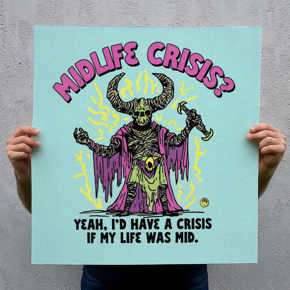 a person holding up a sign with a cartoon-style illustration of a character with horns and a skull-like face, wearing a purple cape and holding a sword. The sign has the text "MIDLIFE CRISIS?" and "YEAH, I'D HAVE A CRISIS IF MY LIFE WAS MID."