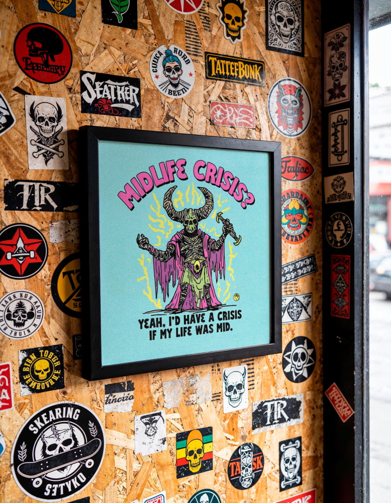 a wall covered in various stickers, patches, and graffiti, with a framed poster hanging on it. The poster has a humorous and artistic design, with the text "MIDLIFE CRISIS? YEAH, I'D HAVE A CRISIS IF MY LIFE WAS MID.
