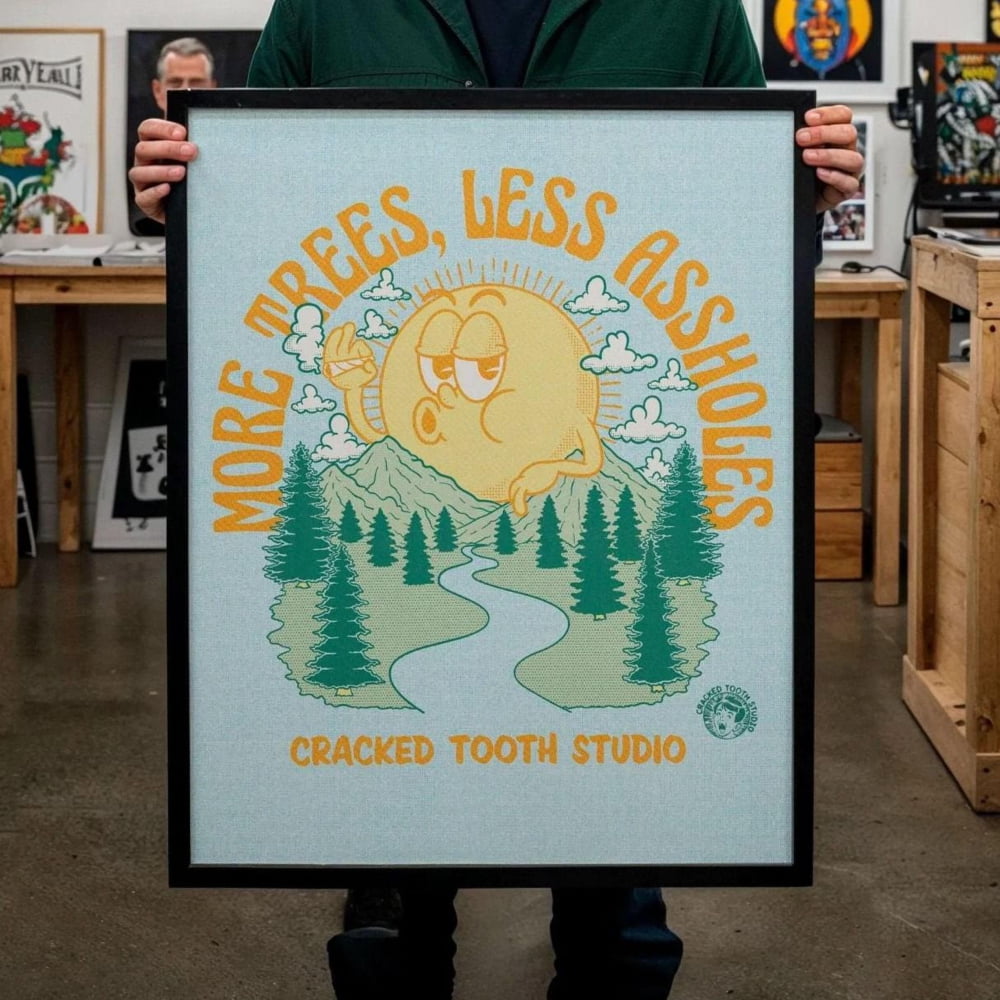 A man is holding up a framed poster with a cartoon-style illustration of a tree and a sun. The poster has the text "More trees, less garbage" and "Cracked Tooth Studio" printed on it.