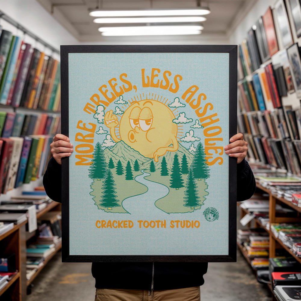 A person is holding up a framed poster with a cartoon sun and trees, featuring the text "MORE TREES, LESS ASSHOLES" and "CRACKED TOOTH STUDIO".