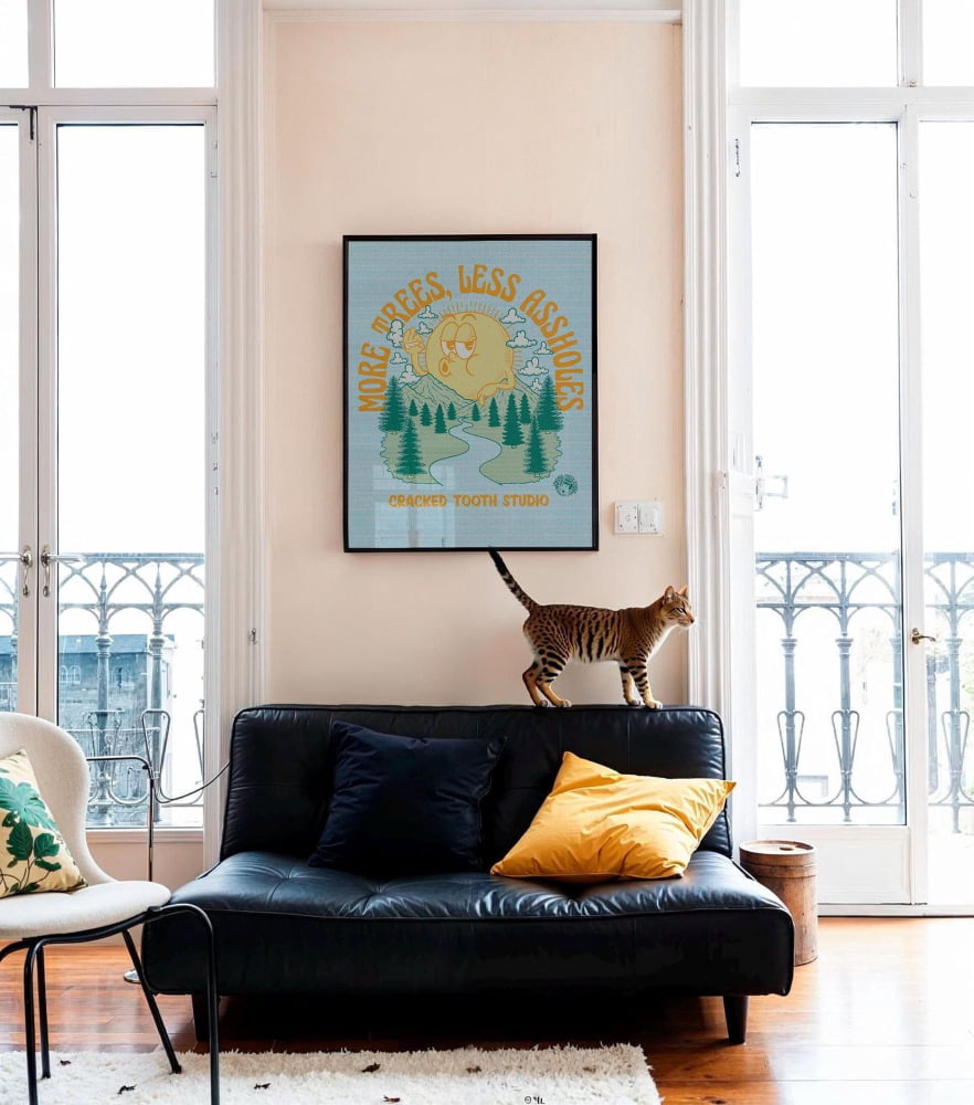 a cat sitting on a black leather sofa with yellow pillows, and a framed poster with a cartoon cat on the wall above the sofa.
