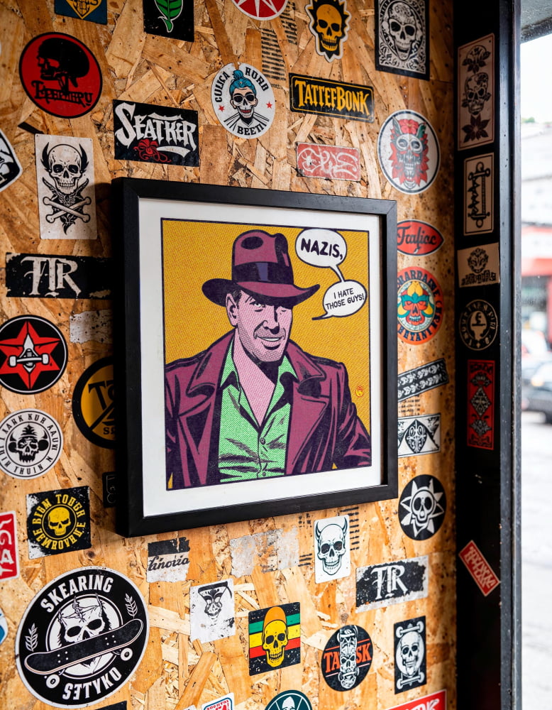a framed illustration of a man wearing a hat, with the caption "NAZIS, I hate those guys" displayed below the portrait. The wall behind the framed image is covered in various stickers, posters, and other graphic designs, creating a visually busy and eclectic backdrop for the artwork.