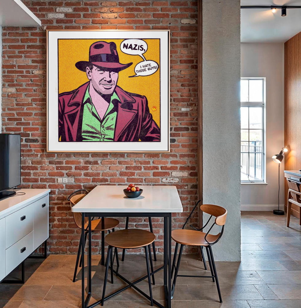 a dining area with a table and chairs, a large framed pop art poster of a man wearing a fedora and a suit, and a brick wall in the background.