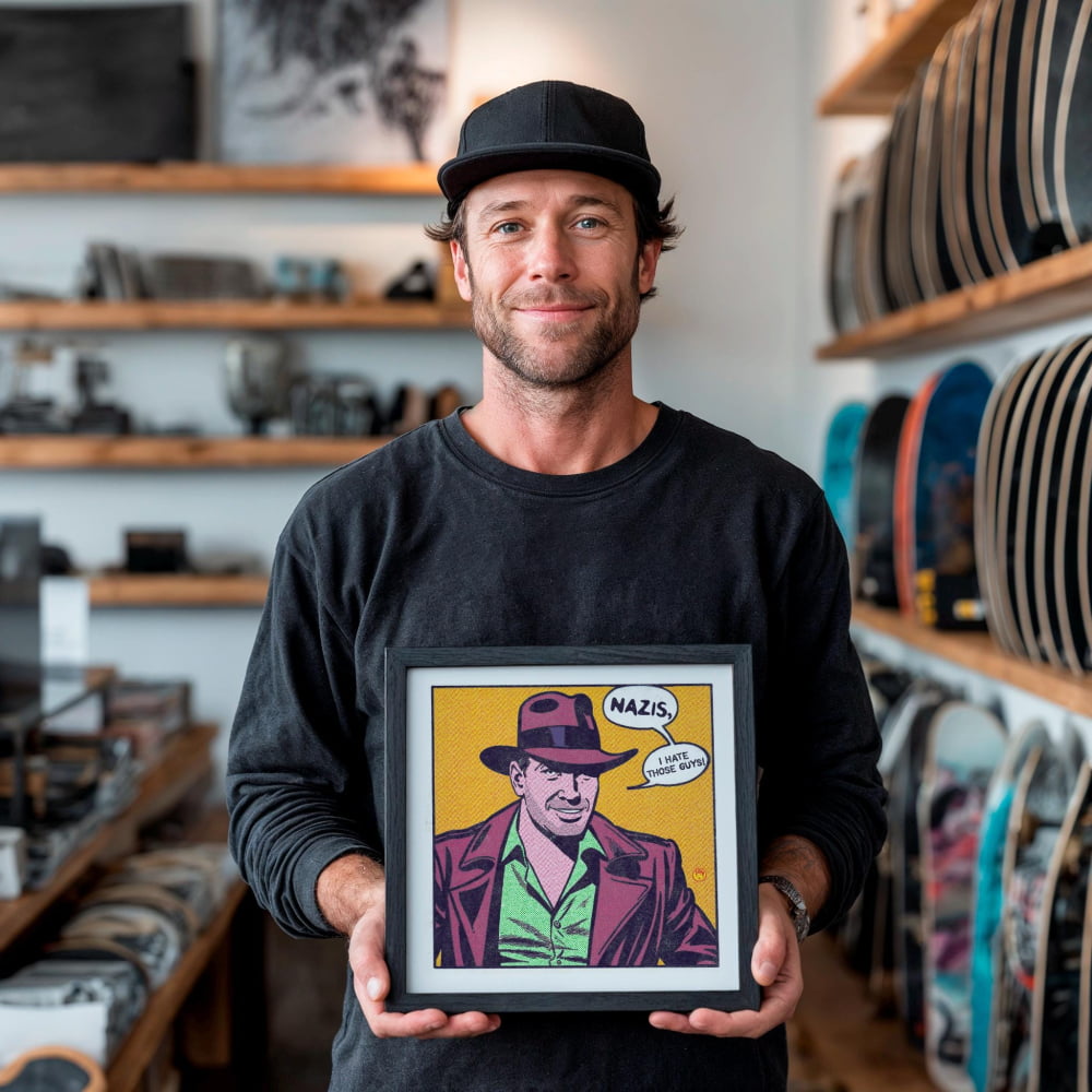 A man is holding a framed comic-style illustration of a man wearing a fedora and a suit, with the caption "NAZIS, I'M NOT HATEFUL OF THOSE THOSE YOU".