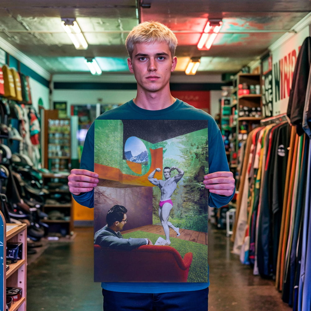 A young man stands in a clothing store, holding up a framed print of a surreal, surreal painting depicting a man in a chair with a woman in the background.