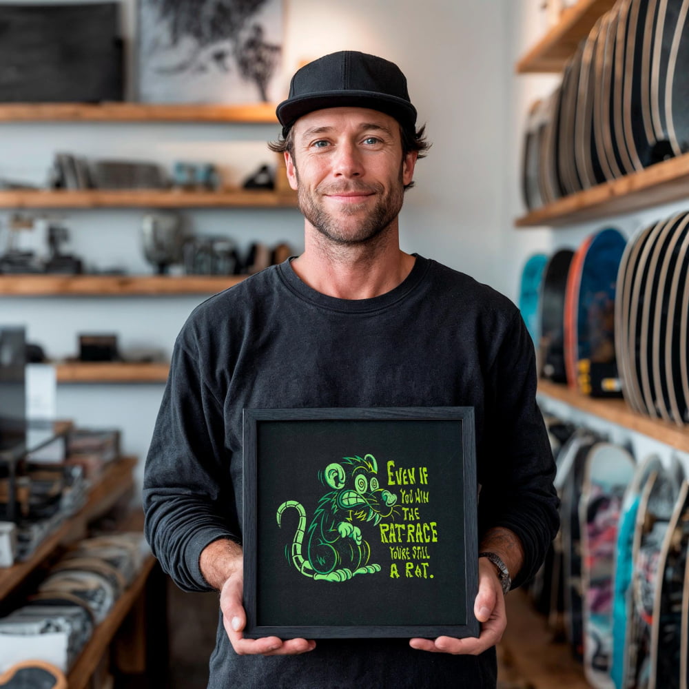 A man is holding a framed picture with a cartoon rat on it, standing in a room with shelves of skateboards in the background.