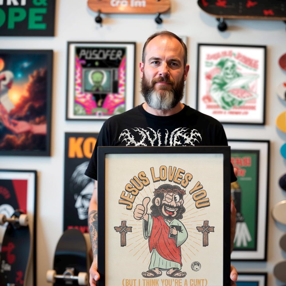 A man with a beard is holding up a framed poster with a cartoon image of Jesus and the text "JESUS LOVES YOU (BUT I THINK YOU'RE A CUT)"