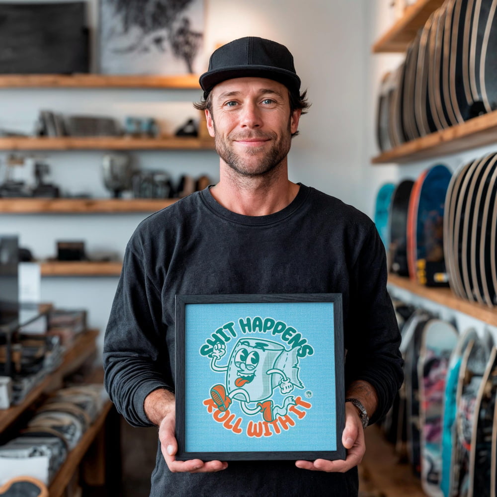 A man is holding a framed picture with a cartoon character on it, which says "SHIT HAPPENS ROLL WITH IT".