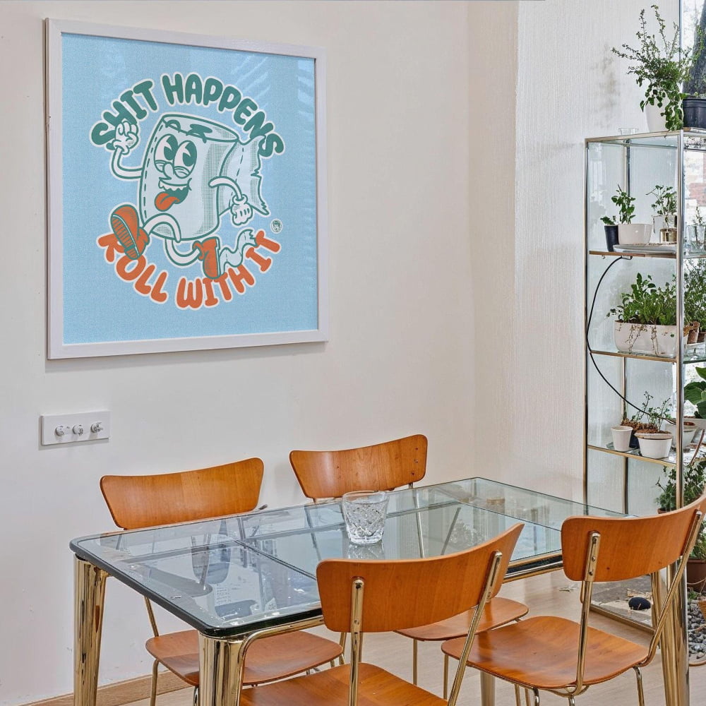 a dining room with a glass table and four wooden chairs. Above the table, there is a framed poster with a cartoon character and the text "SHIT HAPPENS ROLL WITH IT".