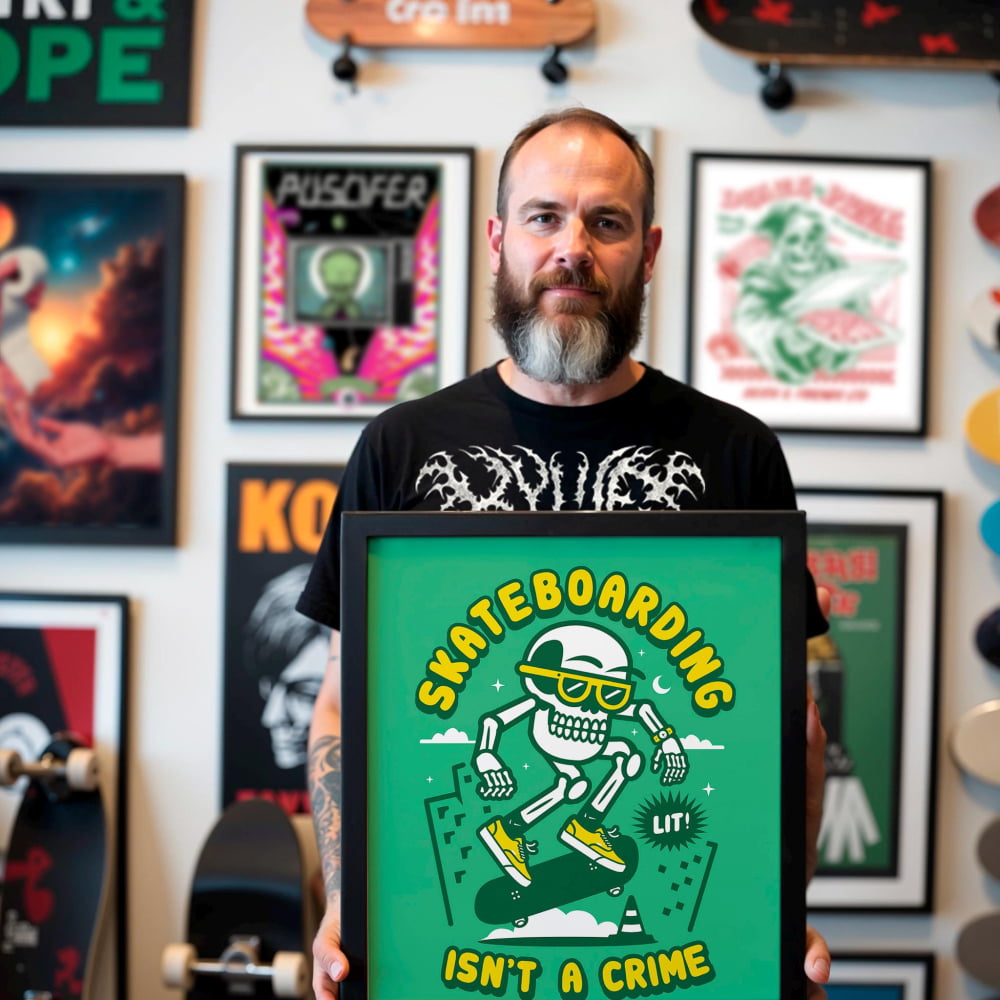 A man with a beard is holding a framed poster that features a cartoon skeleton skateboarding and the text "Skateboarding isn't a crime".
