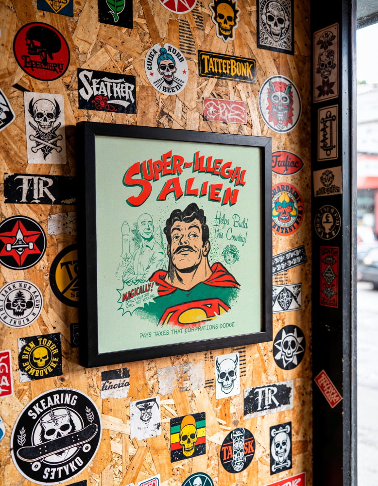 a wall covered in various stickers, posters, and graffiti, including a framed poster with a cartoon character and the text "Super-Illegal Alien".