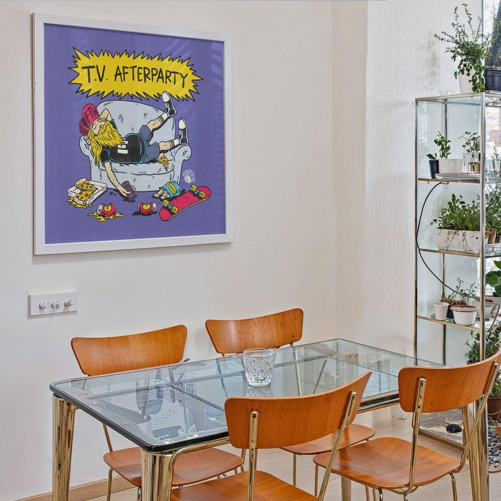a dining room with a glass table and four wooden chairs. Above the table, there is a framed cartoon-style illustration of a person sitting on a couch with a skateboard, surrounded by various objects.