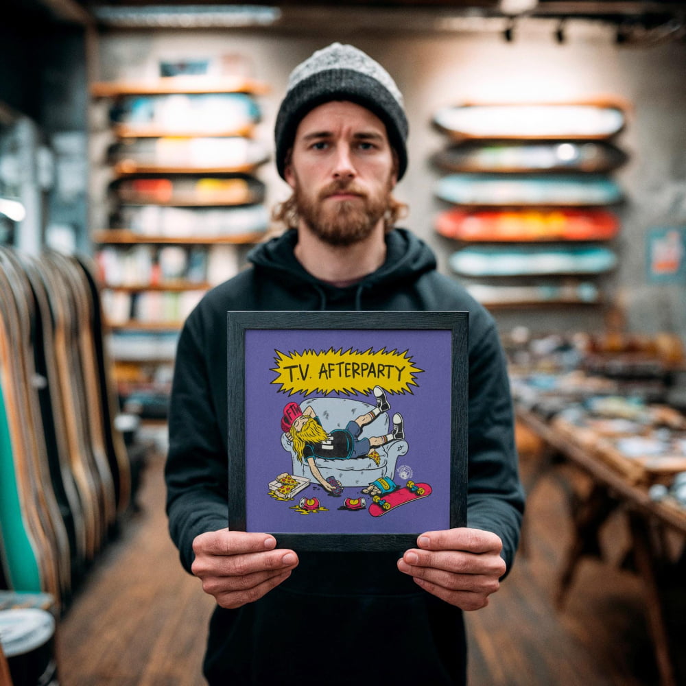 A man holding a framed poster with a cartoon illustration of a person sitting on a couch with a skateboard in their hand.