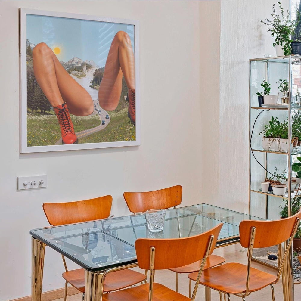 a dining room with a glass table and four wooden chairs. Above the table, there is a framed artwork depicting a woman's legs in a surreal landscape. The room also includes a shelf with potted plants and a glass display case.
