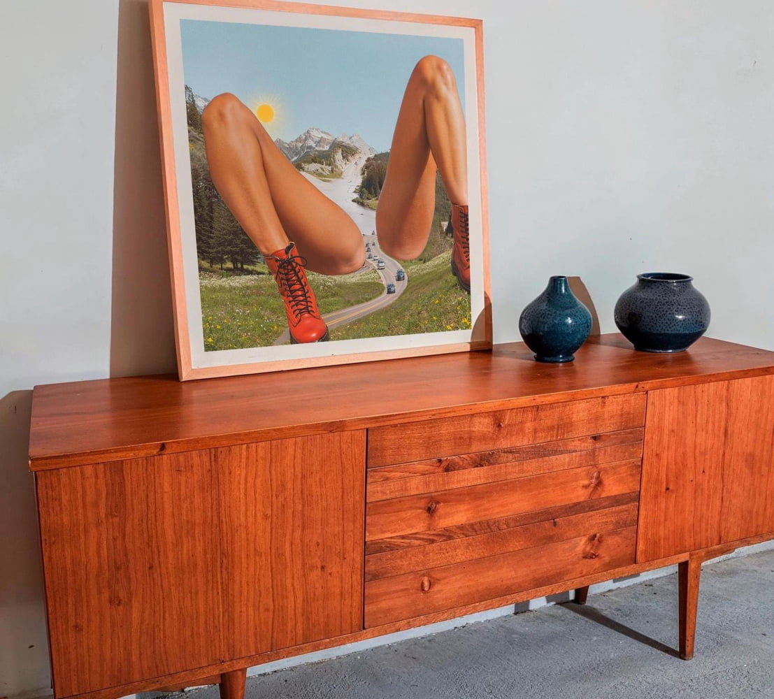 a wooden cabinet with a framed picture of a woman's legs on top of it. The cabinet has two vases on it, one blue and one black.