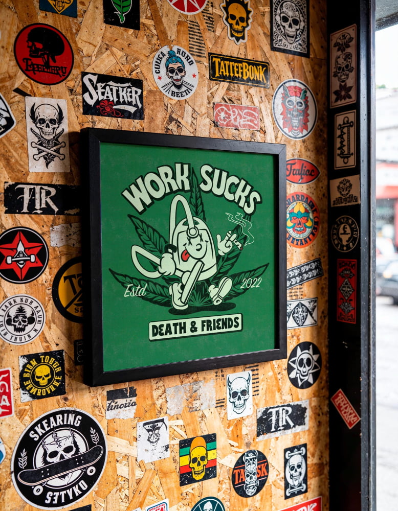 a wall covered in various stickers, patches, and graffiti, including a framed poster with a cartoon character and the text "Work Sucks" and "Death & Friends".